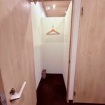 Narrow coat closet with a wooden hanger on a hook and a small trash bin in the corner beneath overhead lights.