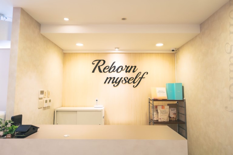 Reception desk in a bright, minimalist lobby with a wood-paneled wall featuring the sign 'Reborn myself' in cursive letters; shelves with turquoise folders and product bags on the right.